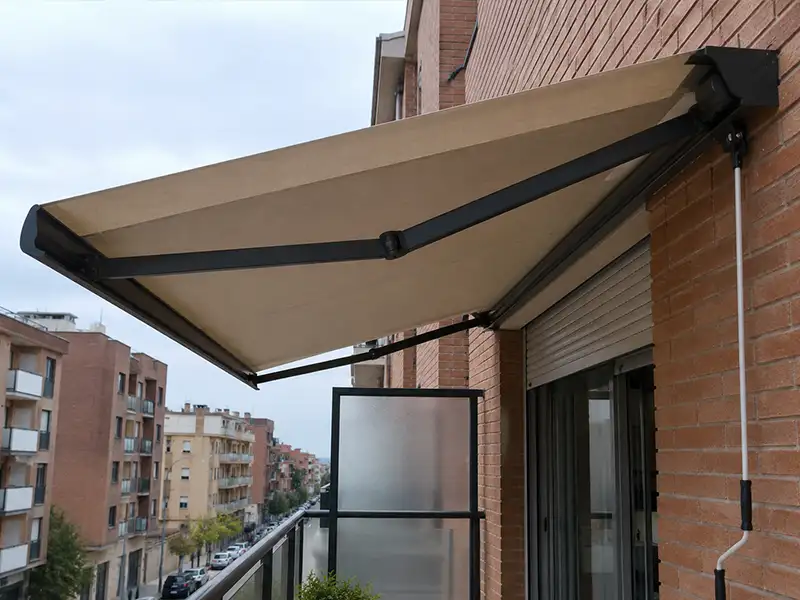 Toldo extensible de brazos articulados con cofre y lona beige instalado en un balcón, proporcionando protección solar para la terraza.