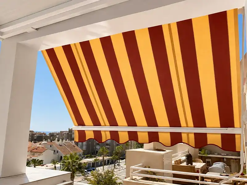 Toldo de punto recto con lona a rayas amarillo y burdeos instalado en balcón de vivienda en Málaga.