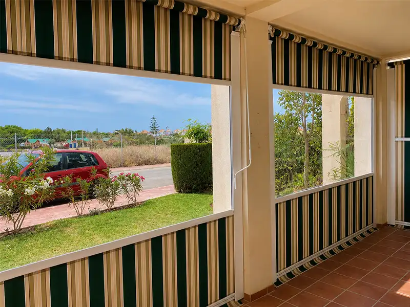 Toldo cortavientos con guías laterales y lona a rayas verde y beige instalado en una terraza para proteger del viento y el sol.